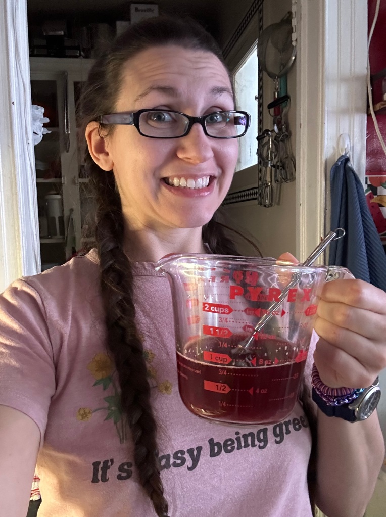 White woman with long brown hair in two braids holding a glass measuring cup with a cup of blood mixed with water and a small whisk